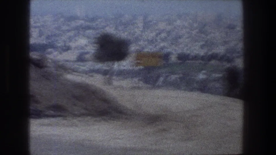Jerusalem hillside buildings pass behind a dark vehicle divider, with a blurred roadside object and hazy 8mm texture.