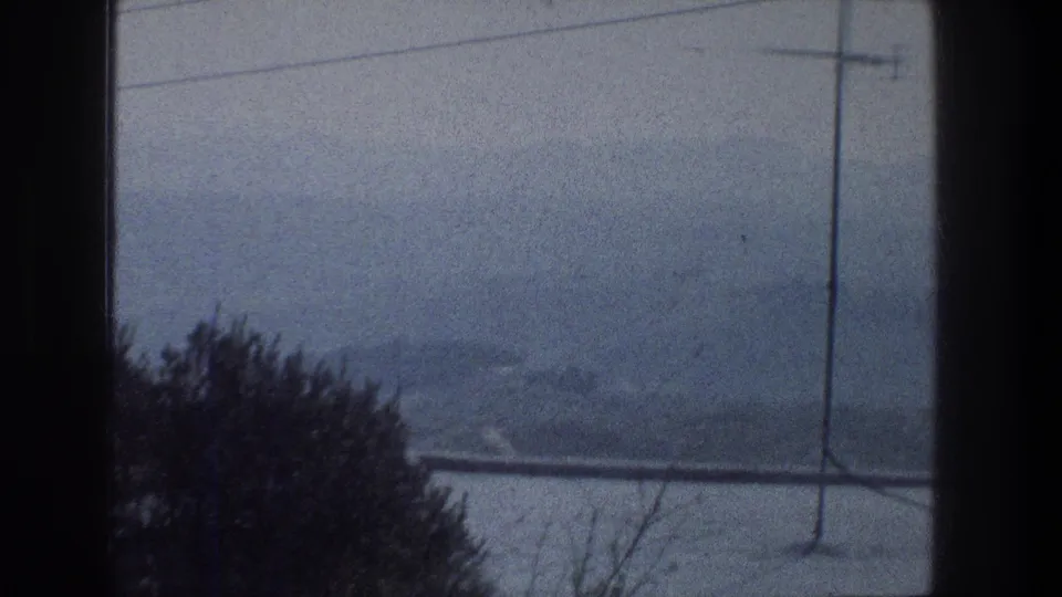 1976 Jerusalem home-movie frame looking through a window toward a hazy hillside, rooftop antenna, wires, and bare branches.