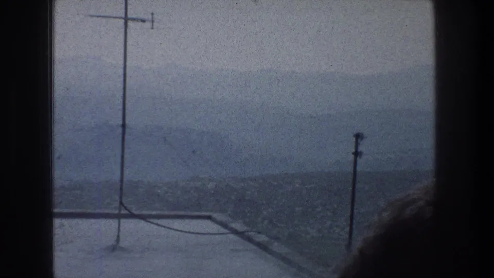 1976 Jerusalem frame with rooftop edge, antenna mast, utility wires, and hazy hillside buildings seen from indoors.