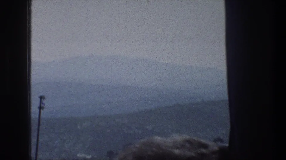 1976 Jerusalem home-movie frame with a soft hazy city and hill view, a pole at left, and a partial head near the window.
