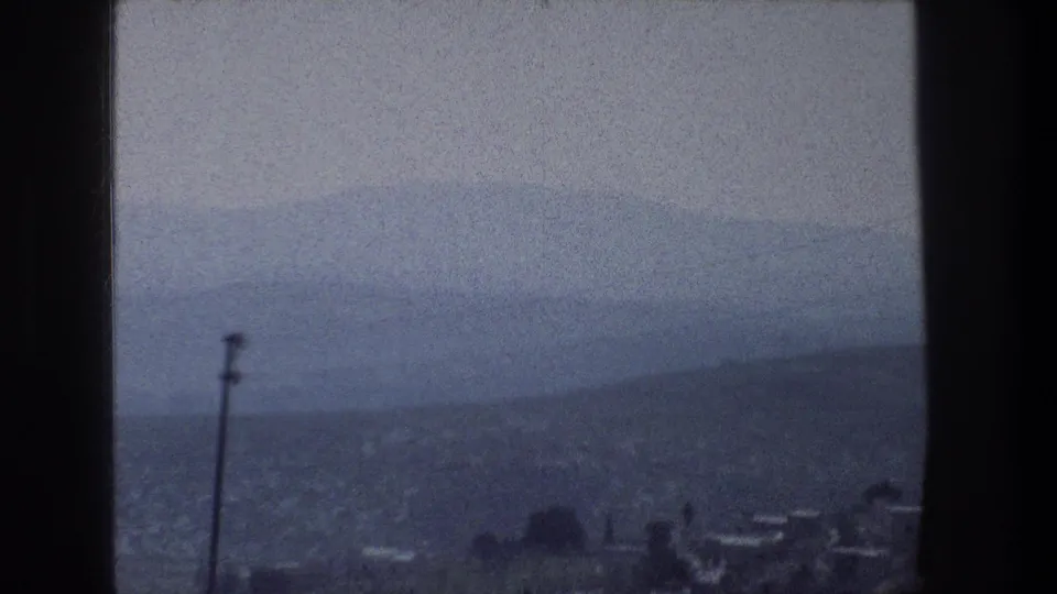 1976 8mm view from a Jerusalem window, showing dark trim, a tall pole, low distant buildings, and hills fading into haze.