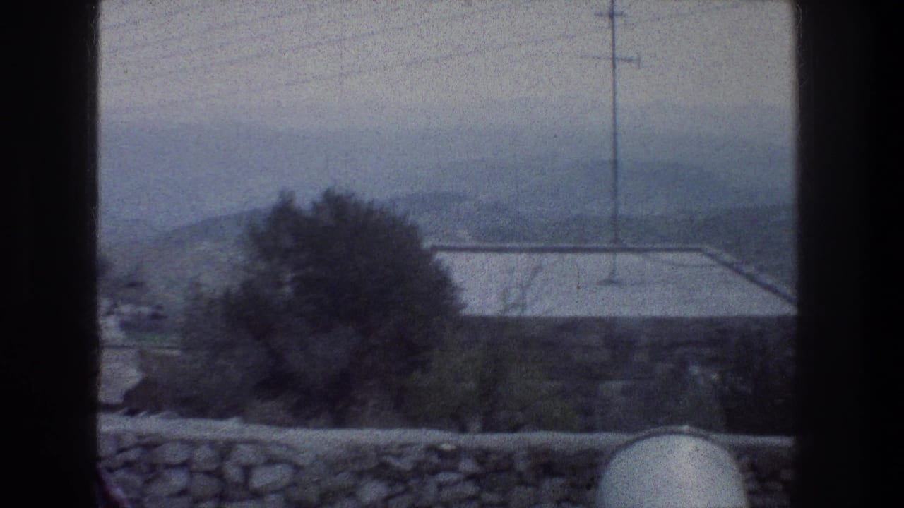 Hazy elevated view past a dark frame, with a stone wall, tree, rooftop, utility pole, wires, and distant hills.