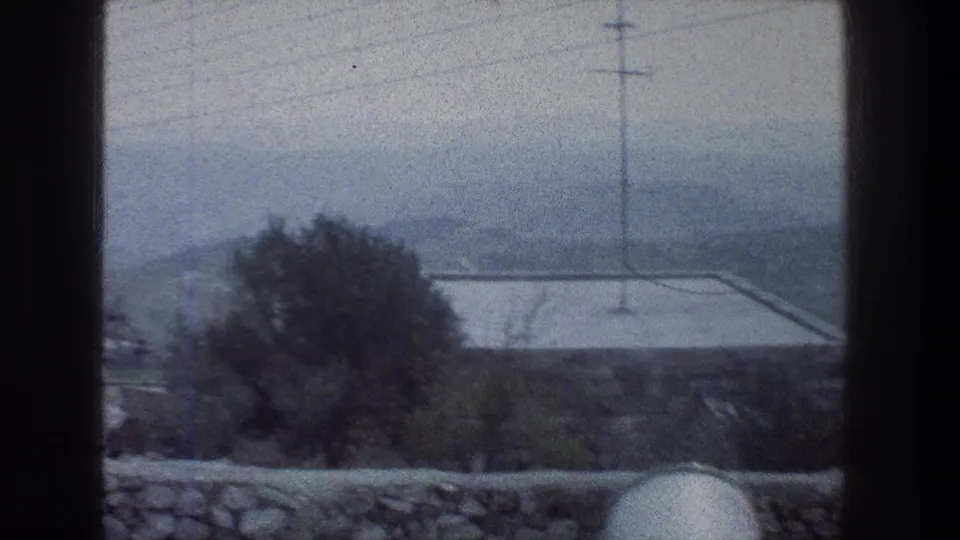 1976 Jerusalem exterior view from above, framed by dark vertical edges, with a tree, stone wall, flat rooftop, utility pole, wires, and hazy hills.