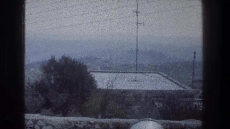 1976 Jerusalem 8mm frame with muted color, showing a foreground stone wall, tree, rooftop, tall utility pole, wires, and distant hazy hills.