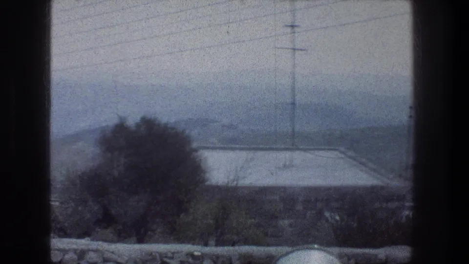 1976 Jerusalem home-movie still looking over a stone wall and tree toward a flat rooftop, utility pole, overhead wires, and misty hills.