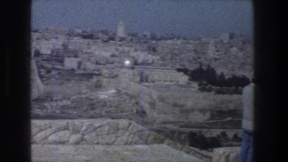 1976 Jerusalem home-movie still showing pale buildings, a long wall, foreground stonework, and dark nearby shapes crossing the view.