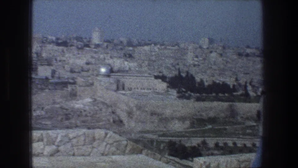1976 Jerusalem frame with stone buildings and walls in the distance and a brief dark foreground person or silhouette near the right edge.