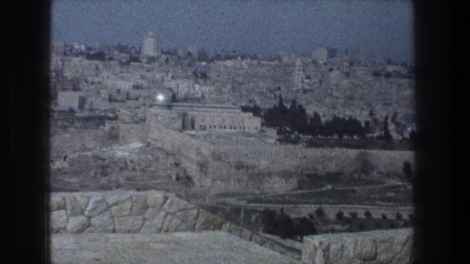 1976 8mm view over Jerusalem, showing a distant domed landmark, stone buildings, trees, and arid terraces under a hazy sky.