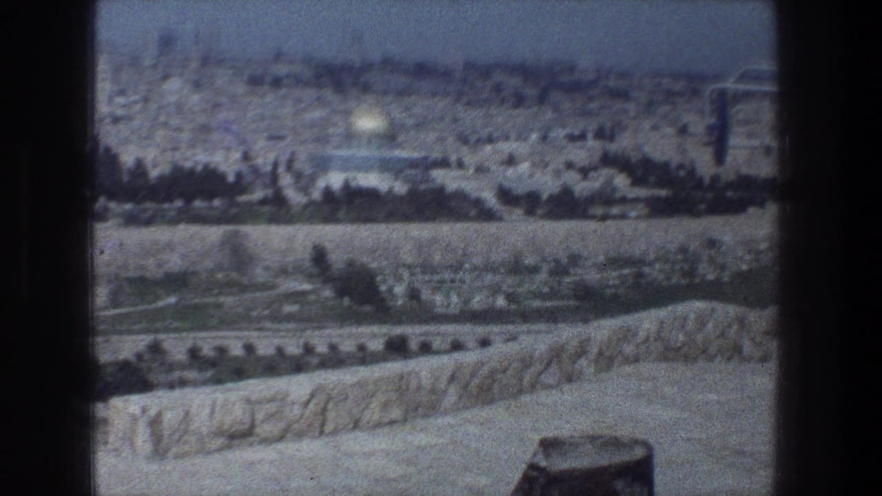 1976 Jerusalem home-movie frame with the Dome of the Rock centered in the distance beyond trees, roads, and pale arid ground.