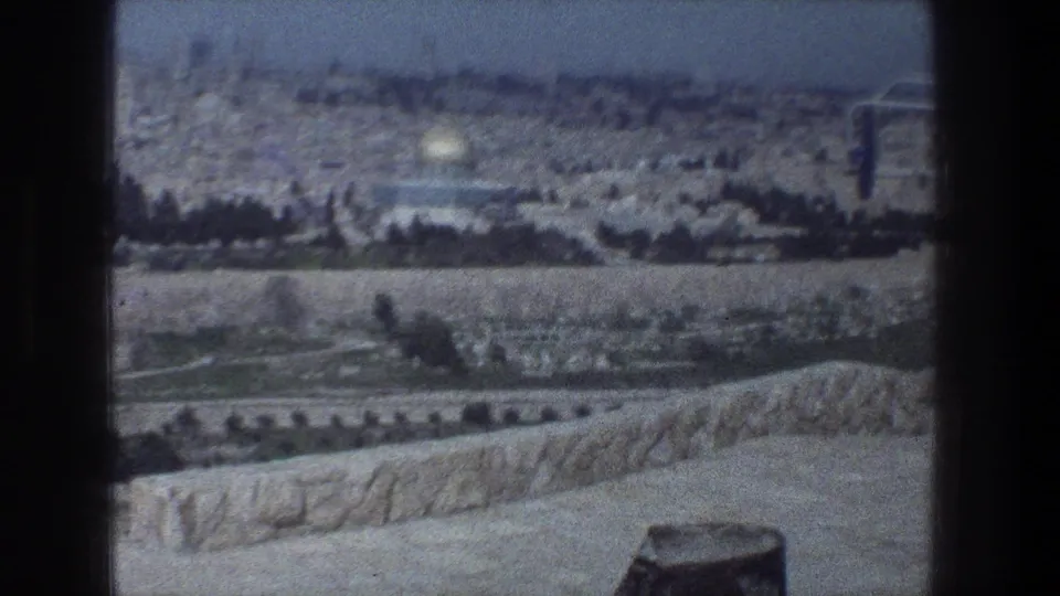 1976 Jerusalem home-movie frame with the Dome of the Rock centered in the distance beyond trees, roads, and pale arid ground.