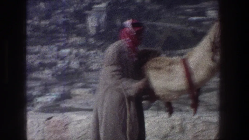 1976 Jerusalem frame showing a handler in a red-and-white keffiyeh guiding a camel through a rocky area.