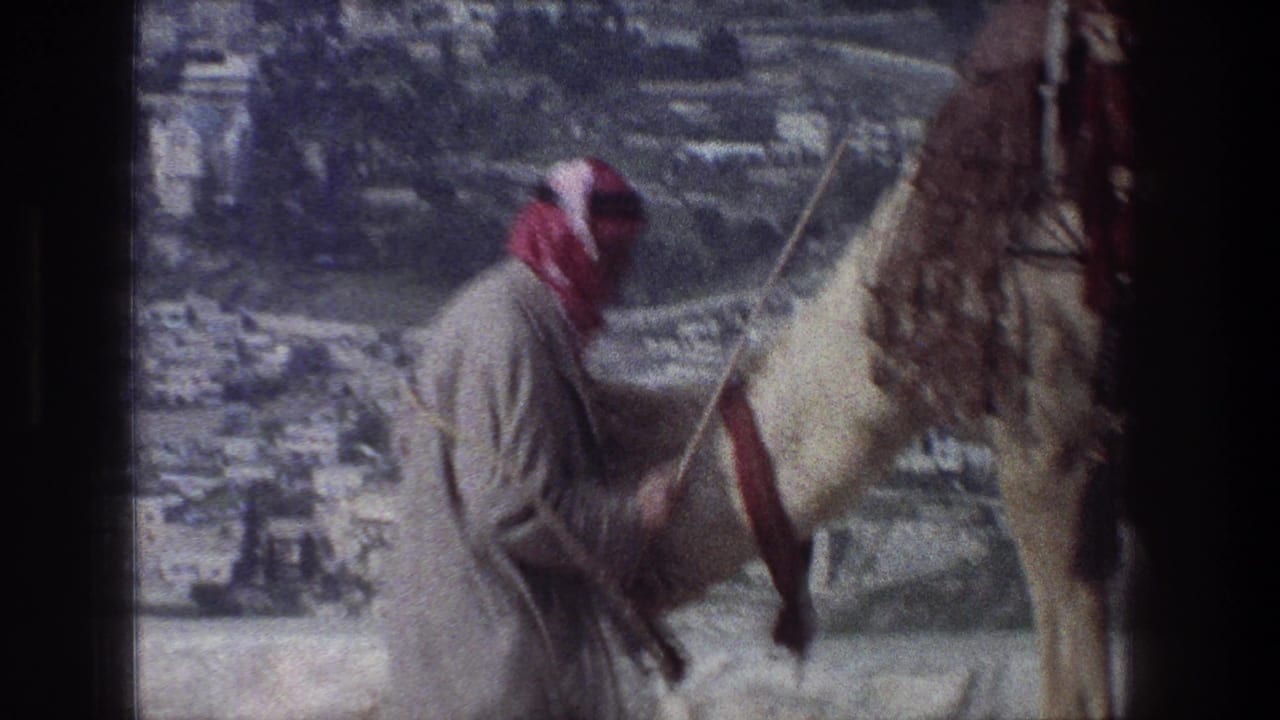 1976 Jerusalem 8mm frame showing a robed man in a red-and-white headscarf guiding a camel near a stone overlook.