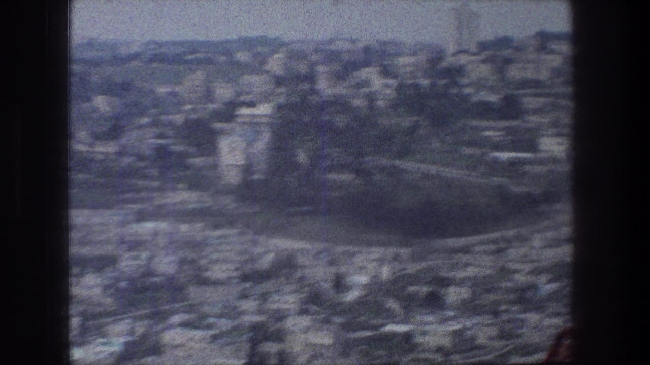 1976 8mm aerial view over Jerusalem, showing dense urban blocks, roads, haze, and a dark projector-like frame edge.