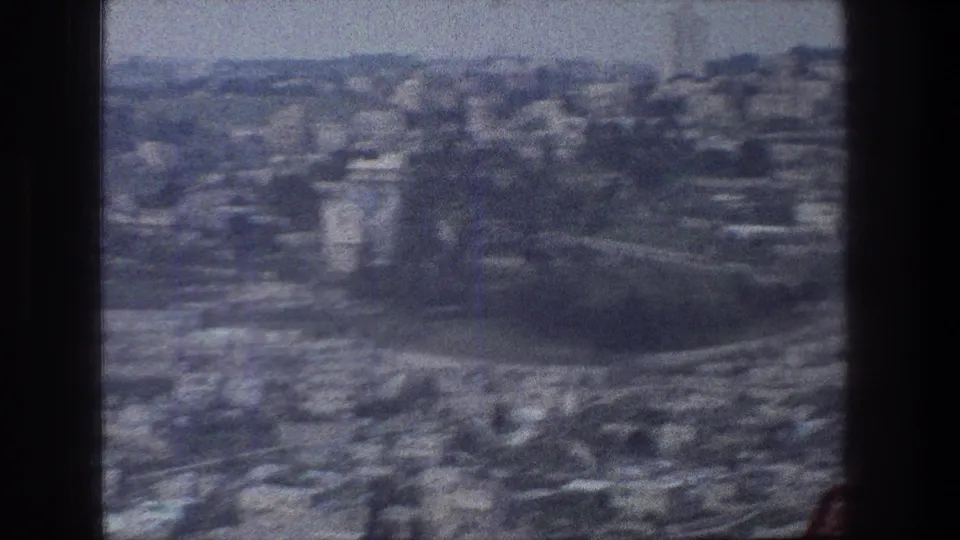 1976 8mm aerial view over Jerusalem, showing dense urban blocks, roads, haze, and a dark projector-like frame edge.