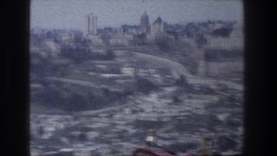 Aerial 8mm frame of Jerusalem with clustered buildings, curving roads, distant towers, and visible film-frame edges.
