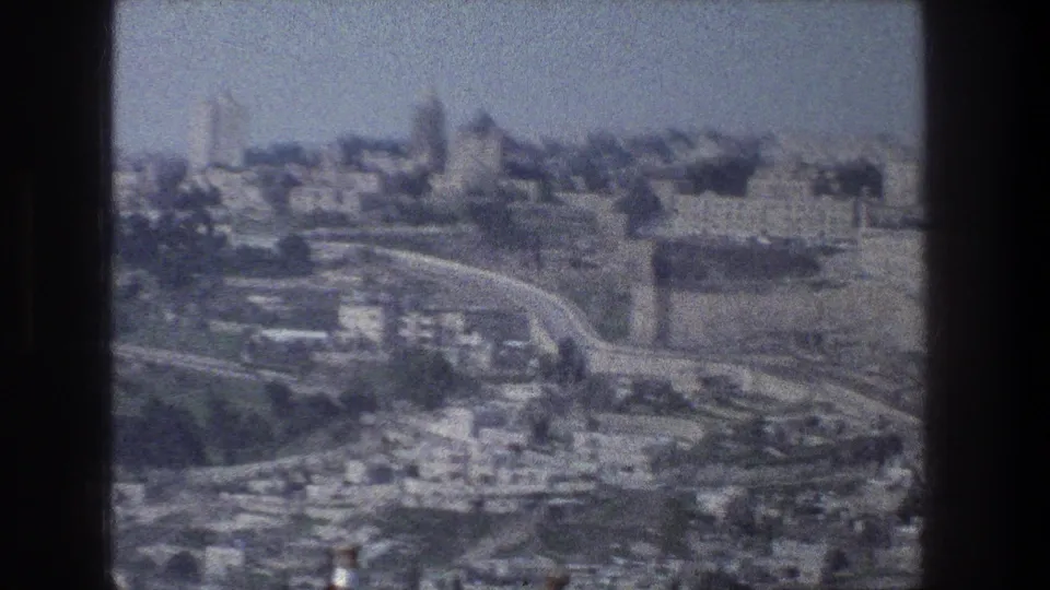 1976 Jerusalem aerial frame showing a built hillside, curving roadways, dense structures, and distant landmark-like forms.