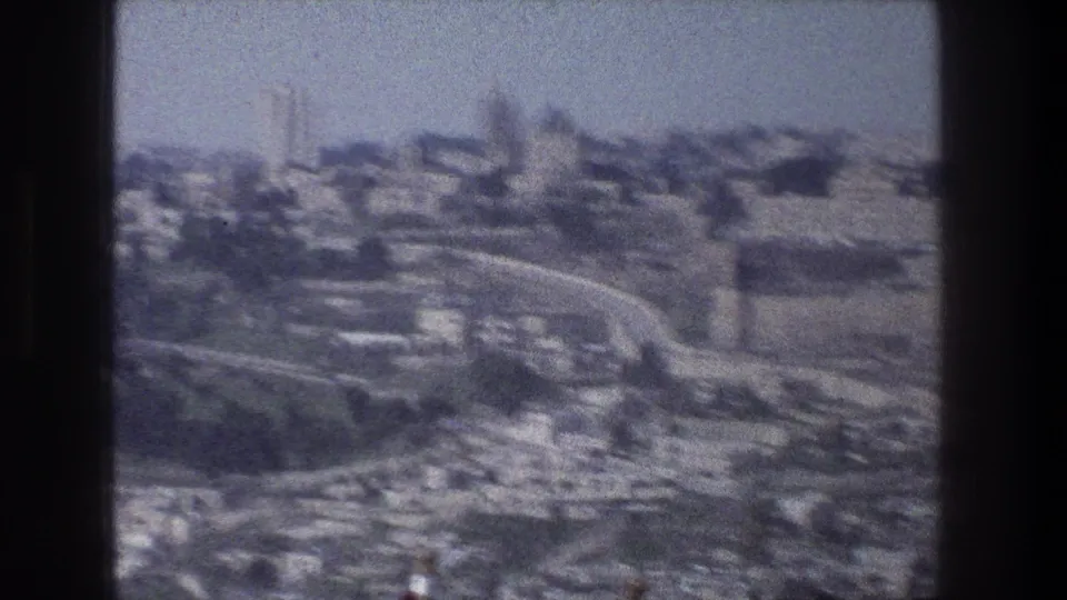 Soft 1976 8mm aerial view of Jerusalem, with clustered buildings, winding roads, distant skyline, and heavy film grain.