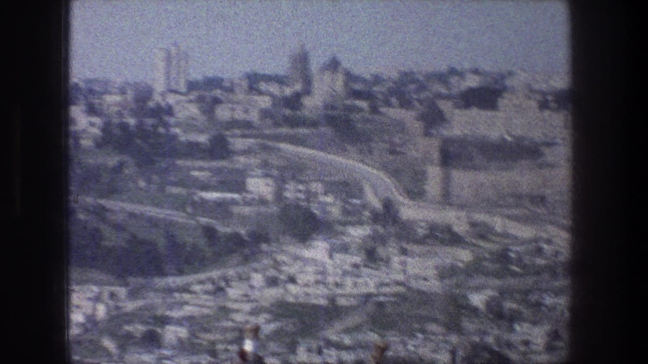1976 Jerusalem home-movie frame showing a distant city view with clustered stone buildings, roads, hillside terrain, and visible film gate edges.