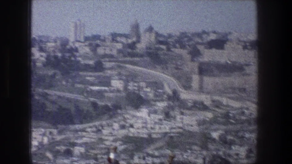 1976 Jerusalem home-movie frame showing a distant city view with clustered stone buildings, roads, hillside terrain, and visible film gate edges.