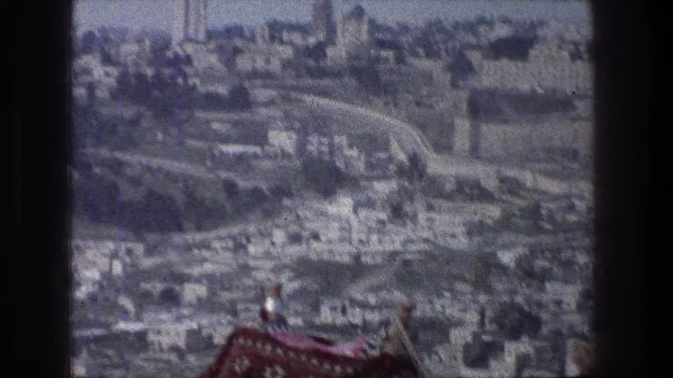 1976 Jerusalem balcony view with dense stone buildings, winding routes, old wall lines, and a red patterned textile visible in the foreground.