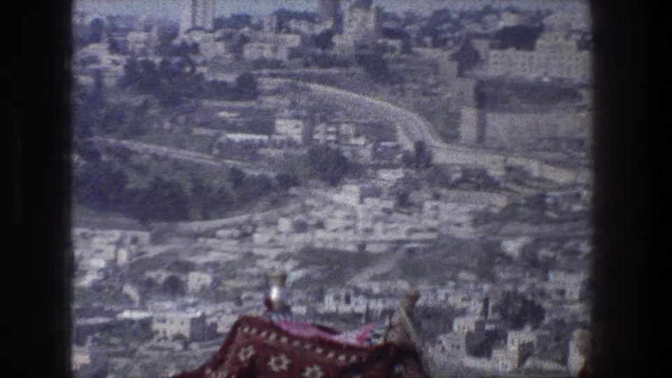 1976 8mm Jerusalem frame showing a soft, grainy urban panorama from a high viewpoint with stone structures and foreground patterned fabric.