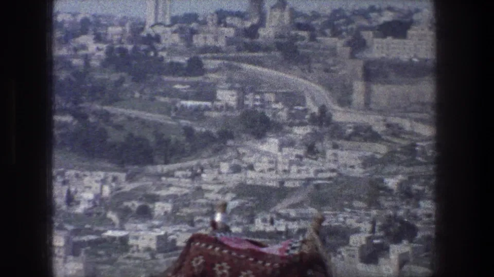 1976 Jerusalem home-movie still with a hazy cityscape, curving roads, dense stone buildings, and a red patterned textile at the bottom edge.