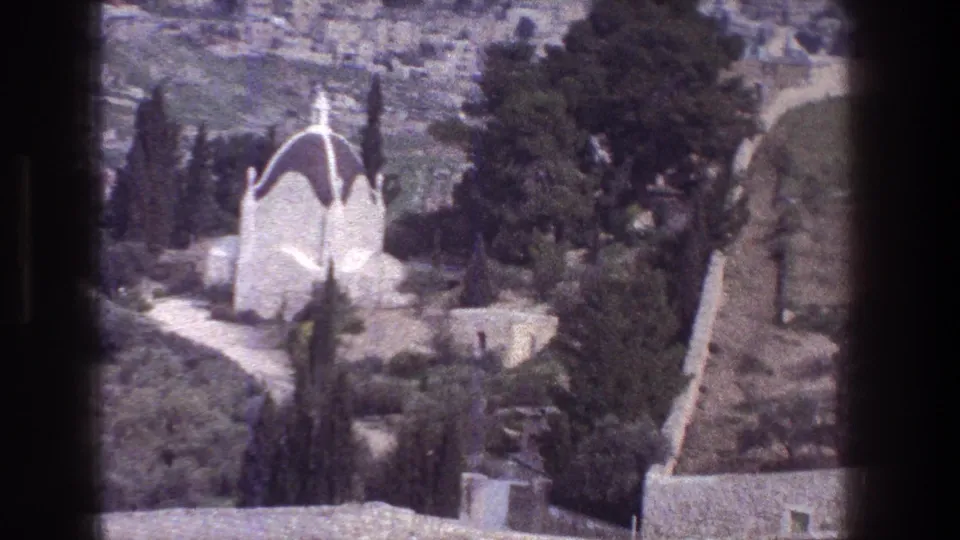 1976 Jerusalem home-movie frame with a white domed building, tall cypress trees, greenery, and stonework in soft focus.