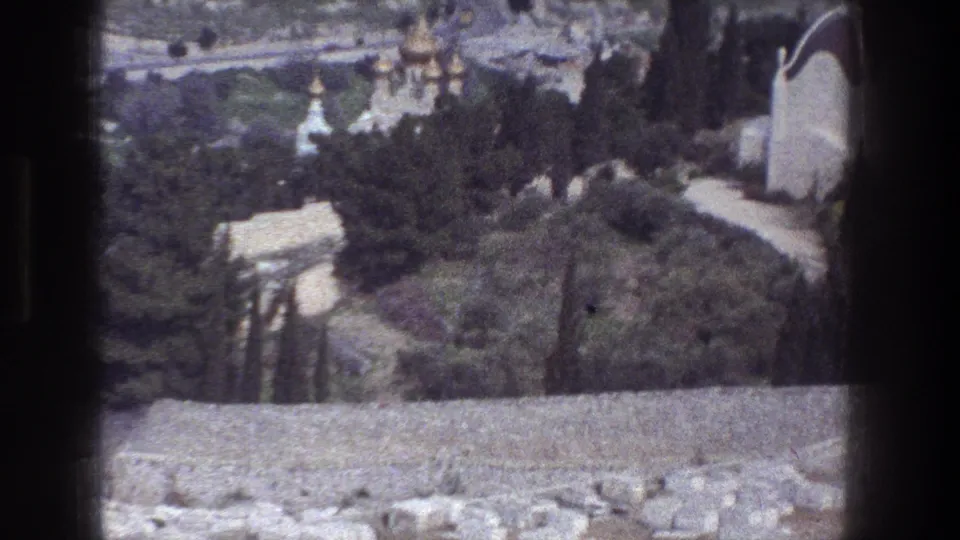 Elevated 1976 Jerusalem home-movie frame showing a stone parapet, dense trees, hillside paths, and distant domed buildings.