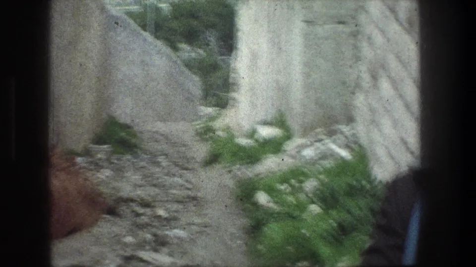 Close visitor-shot 8mm view in Jerusalem showing a stone path, pale wall, shrubs, and part of a person at the frame edge.