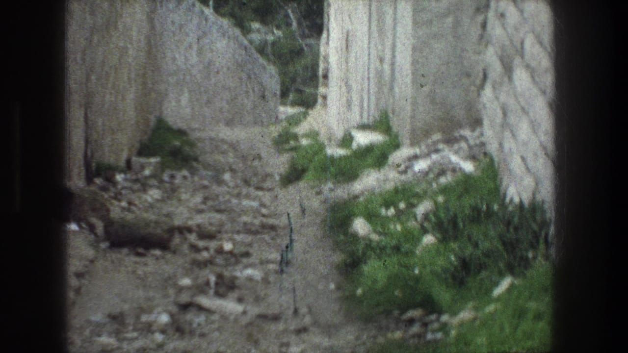 Jerusalem ruin passage in 1976 home-movie footage, with a rough stone wall, scattered rocks, and green growth along the narrow path.