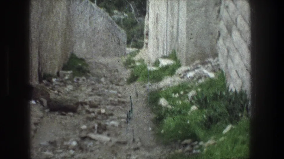 Jerusalem ruin passage in 1976 home-movie footage, with a rough stone wall, scattered rocks, and green growth along the narrow path.