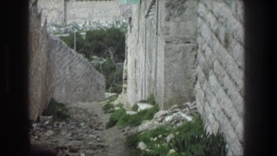 Handheld 8mm view of a Jerusalem stone corridor, with high masonry walls, rubble on the path, and vegetation near an opening ahead.