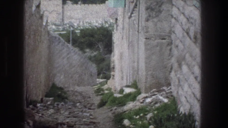 1976 Jerusalem 8mm frame of a narrow ruin path bordered by weathered stonework, scattered debris, and patches of green vegetation.
