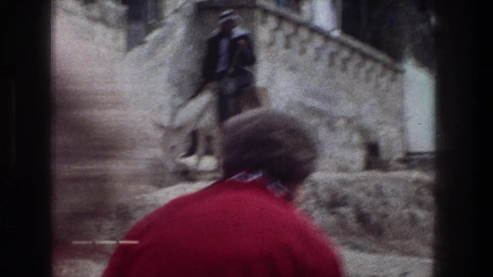 1976 Jerusalem home-movie frame with a person in a red shirt in the foreground and a man walking up stone steps in the distance.