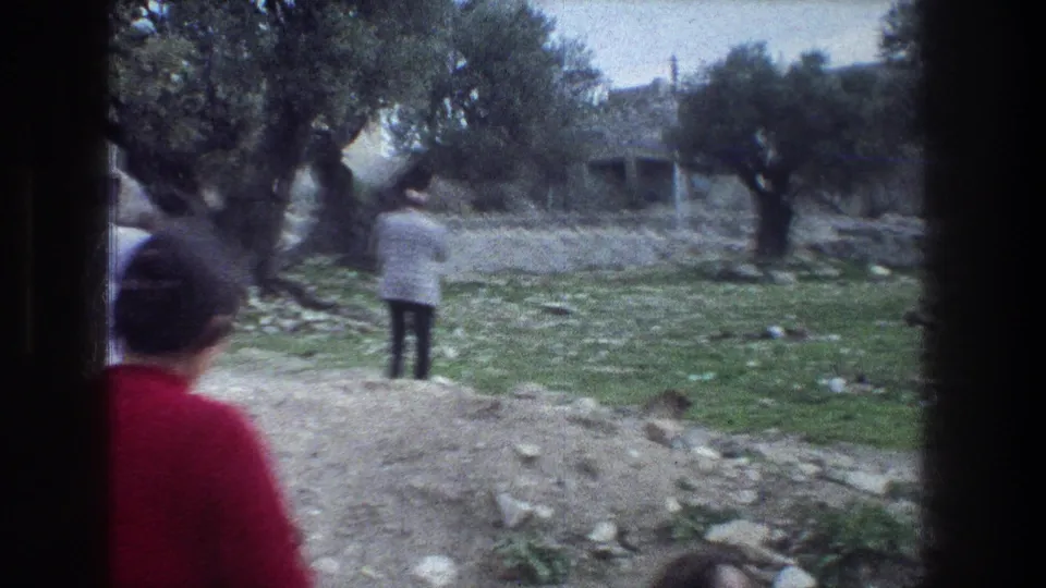 1976 Jerusalem home-movie frame of a man walking away among olive trees, stone walls, and uneven rocky ground.