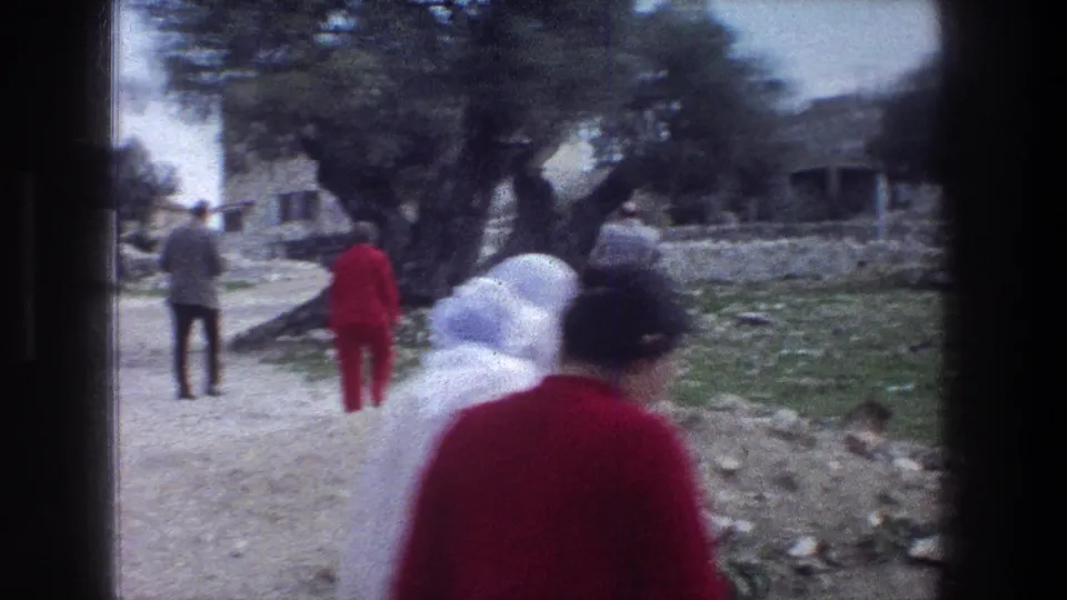 1976 Jerusalem 8mm frame showing several people walking near a rocky field, low stone walls, trees, and distant buildings.
