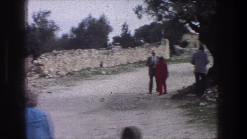 A 1976 Jerusalem 8mm frame shows adults talking or pausing on a path bordered by old trees and stone walls.