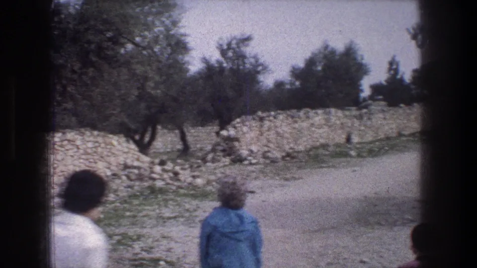 1976 Jerusalem home-movie frame with adults at the edge of a path, a blue jacket in the foreground, and trees behind a stone wall.