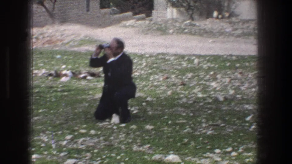 1976 Jerusalem home-movie frame showing an adult man kneeling in a rocky grassy field while using a camera.