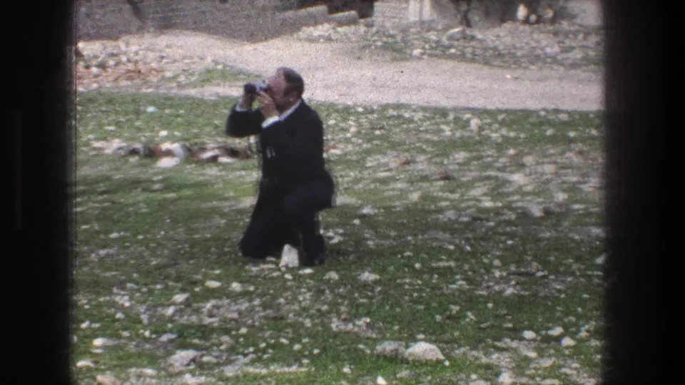 1976 Jerusalem 8mm frame of an adult man kneeling with a camera in a rocky field bordered by low stone walls.