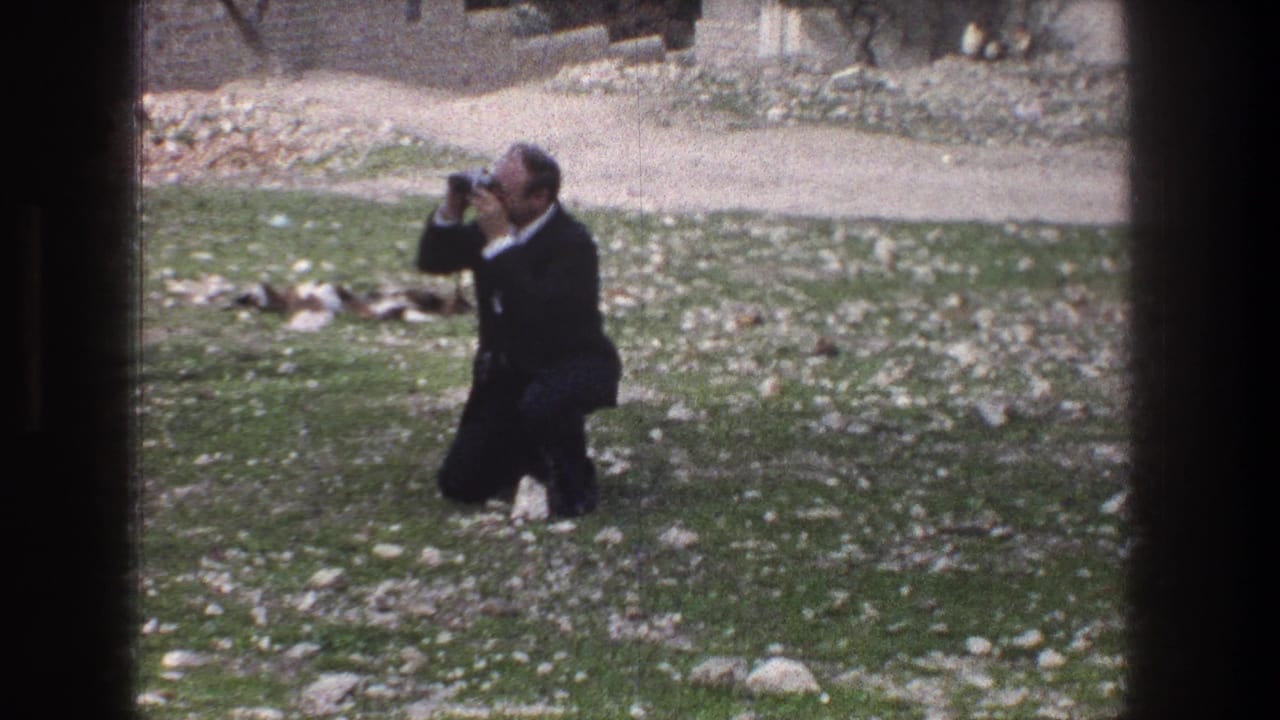 Man in a dark suit crouching on grass while raising a camera.