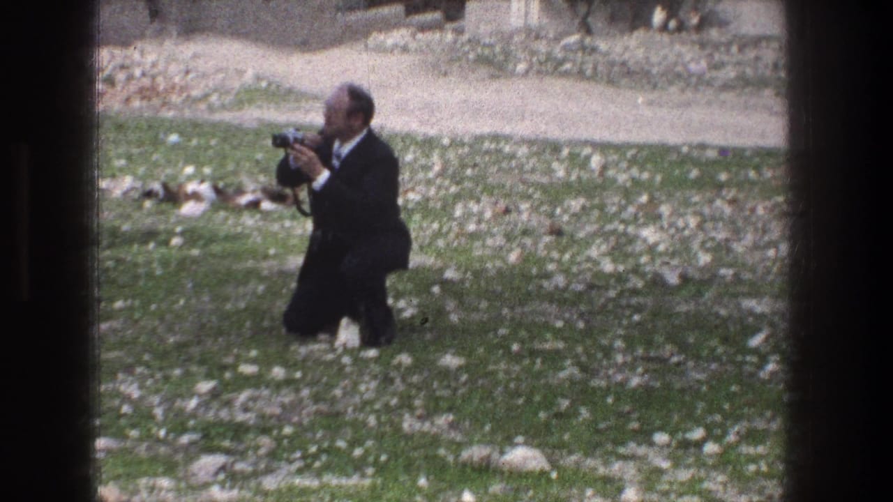 A man in a dark suit crouches on grass in Jerusalem, aiming a vintage still camera across the open area.