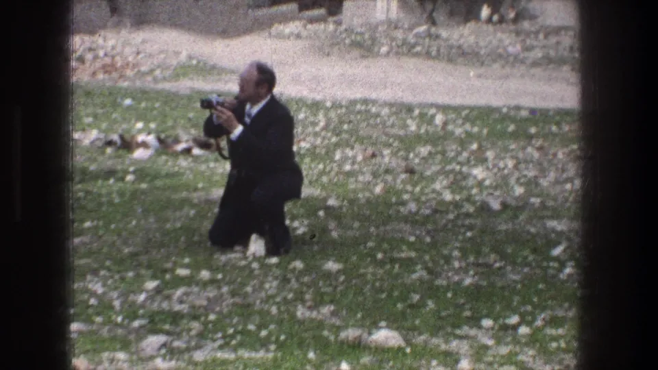 A man in a dark suit crouches on grass in Jerusalem, aiming a vintage still camera across the open area.