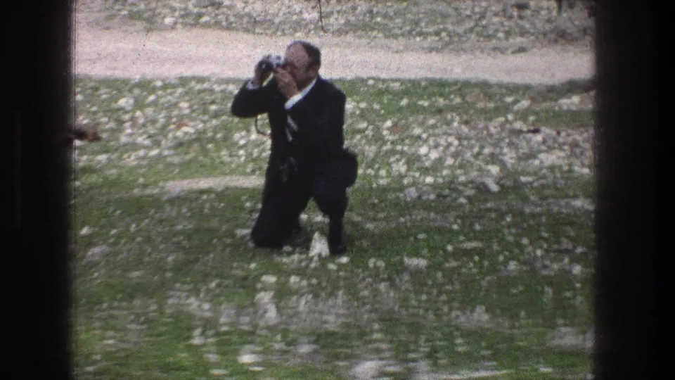 The 8mm frame shows the suited man crouched low on a rocky lawn, holding the camera to his eye in the Jerusalem setting.