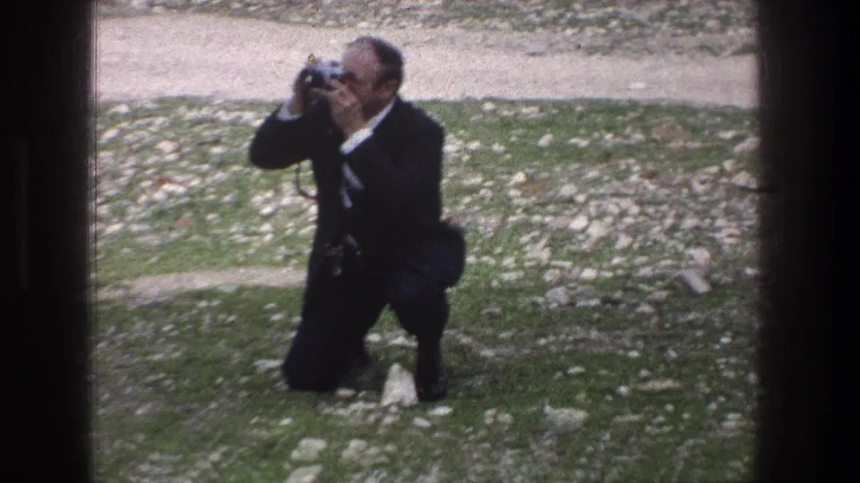 A closer 1976 Jerusalem frame shows the man crouched on the uneven lawn, camera pressed to his face as he takes a photograph.