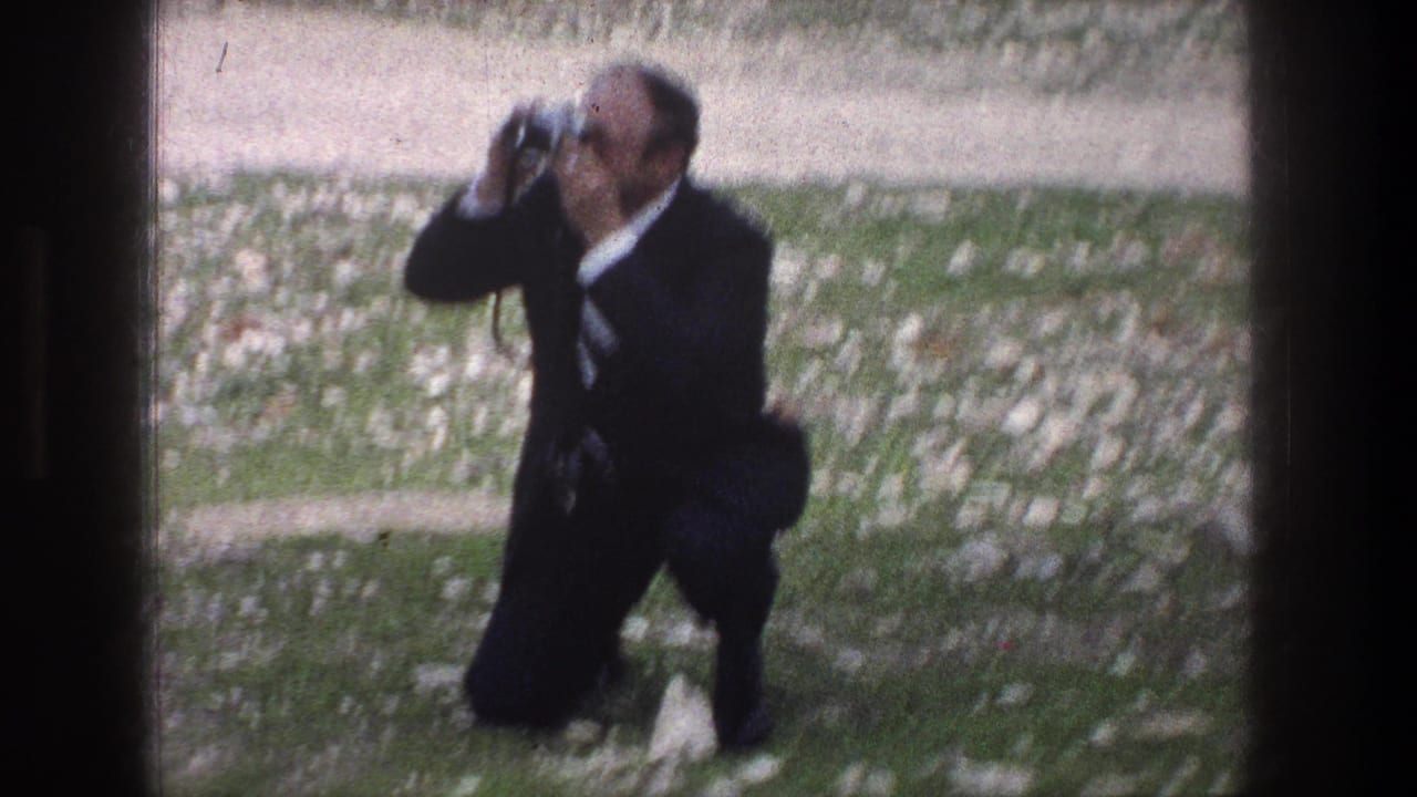 In 1976 Jerusalem footage, a suited adult kneels on rocky grass and raises a camera to photograph something off frame.