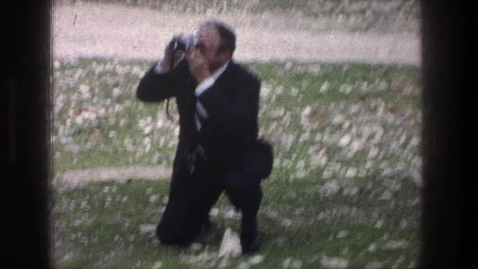 In 1976 Jerusalem footage, a suited adult kneels on rocky grass and raises a camera to photograph something off frame.
