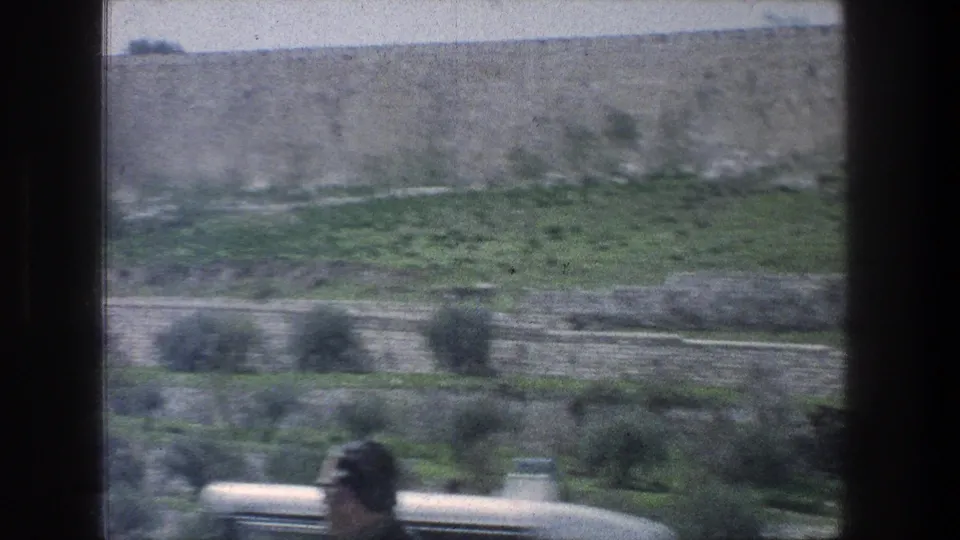 1976 Jerusalem roadside landscape with stone terraces, green vegetation, and a brief blurred foreground figure wearing glasses.