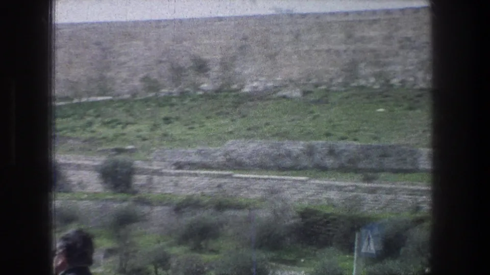 1976 Jerusalem moving-vehicle frame with terraced greenery, stone retaining walls, a blue triangular road sign, and a blurred foreground person.