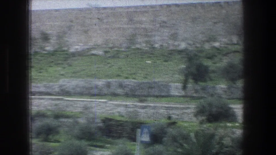 1976 Jerusalem roadside view of layered stone terraces, green vegetation, and a blue triangular road sign near the bottom edge.
